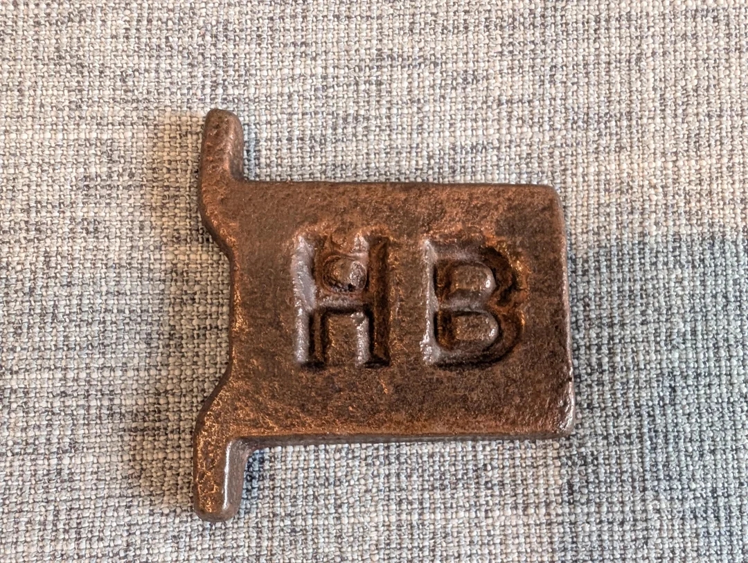 A rustic metal branding iron with "HB" embossed on it, lying on a textured surface