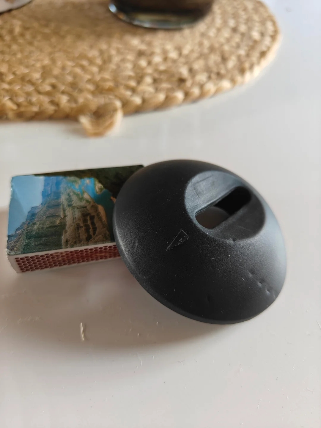 A black item resembling a dome with an opening sits beside a matchbox on a table with a braided mat in the background