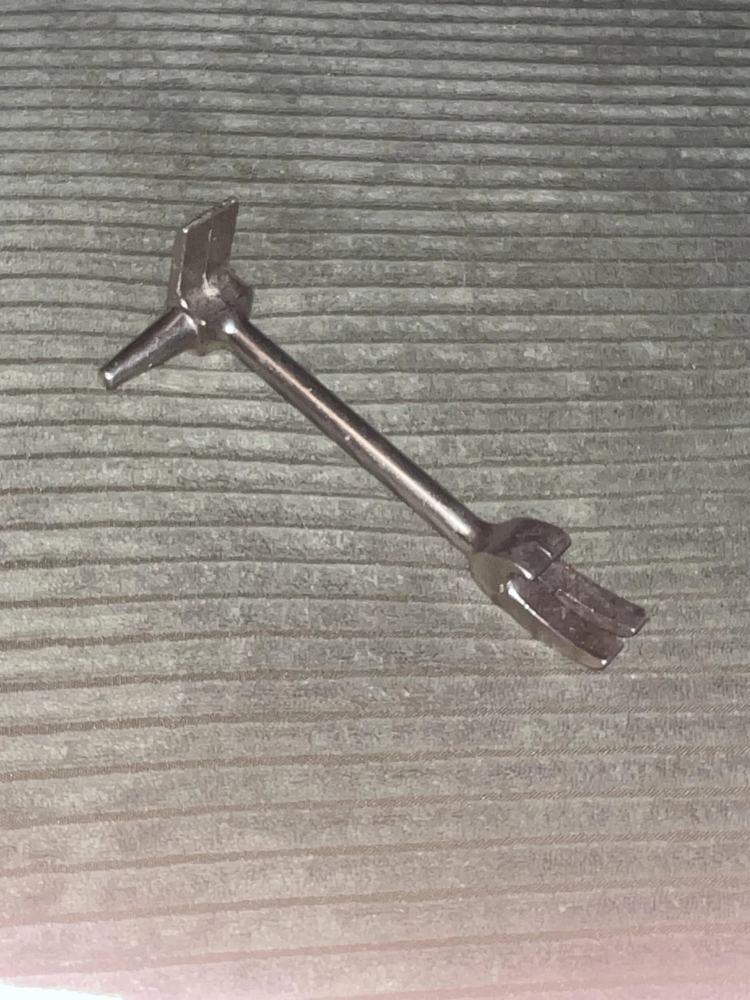 Metal tool resembling a T-shaped wrench lays on a textured surface; purpose and specific function unclear