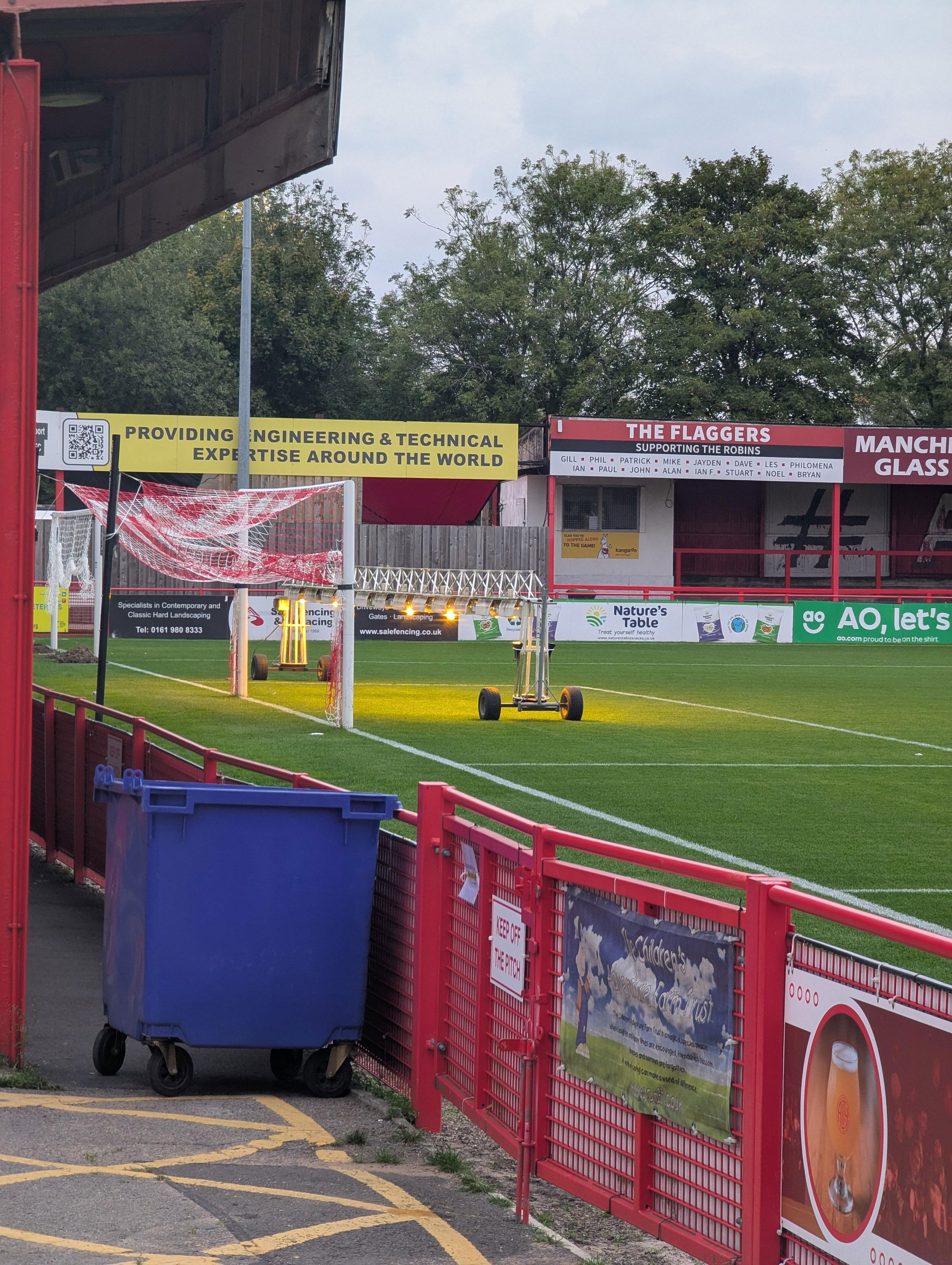 Soccer field with advertising banners in the background, highlighting engineering and technology expertise. A blue recycling bin is visible in the foreground