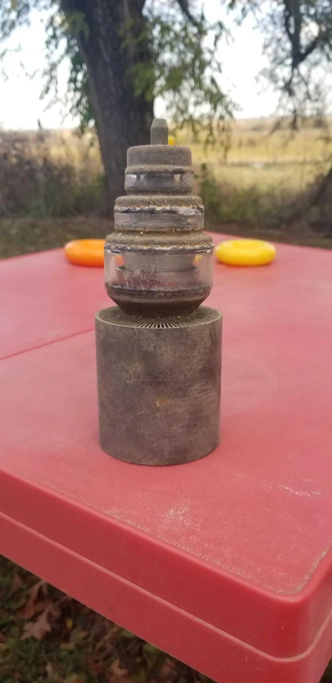 Mysterious metal object with a cylindrical base and layered components on a red table outdoors. Trees and grass are visible in the background