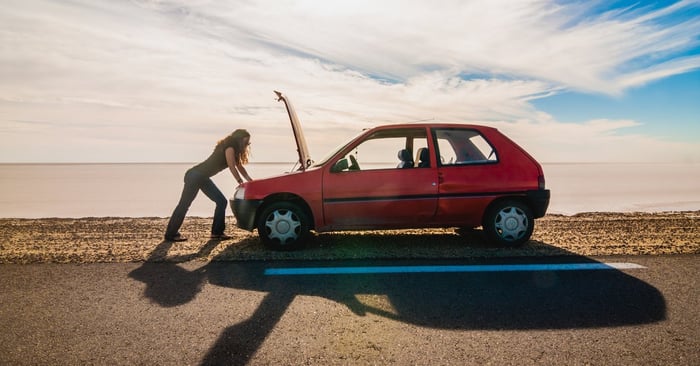 woman standing on the side of the road looking under the hood of her car.