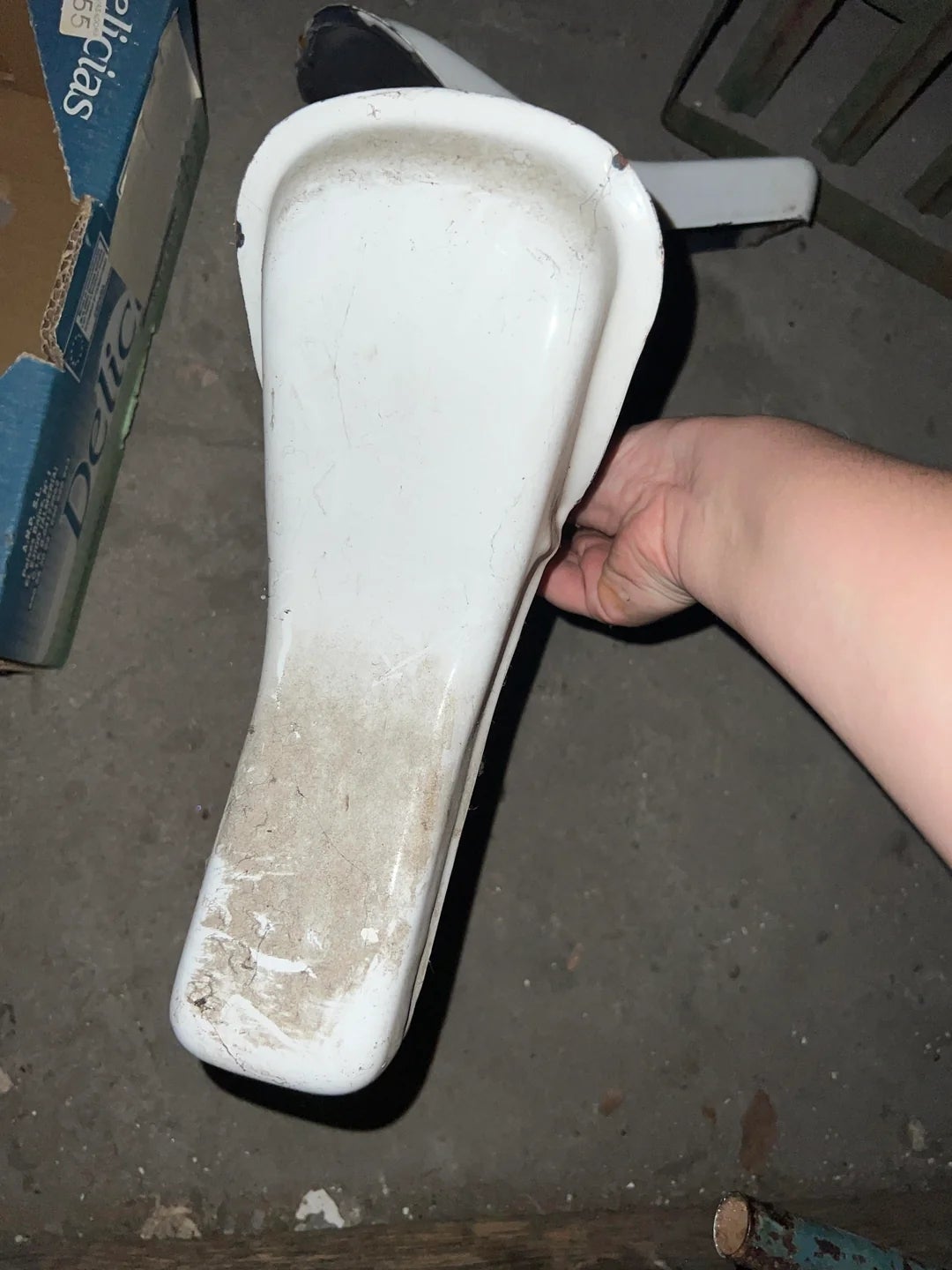 Person holding a white plastic object, possibly a seat, in a cluttered space with boxes and items around