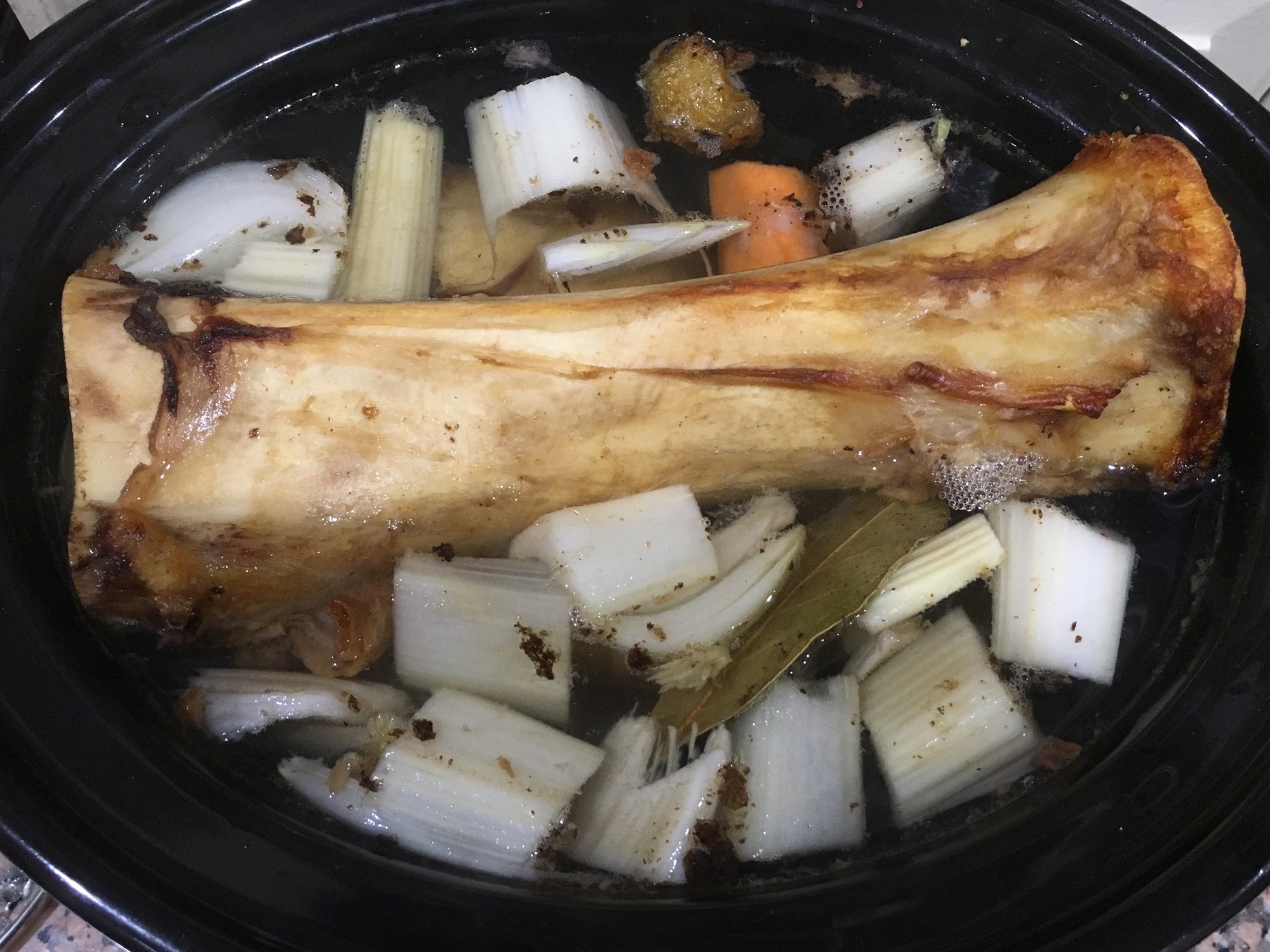 Bone and vegetable broth cooking in a slow cooker with onions, carrots, celery, and bay leaf