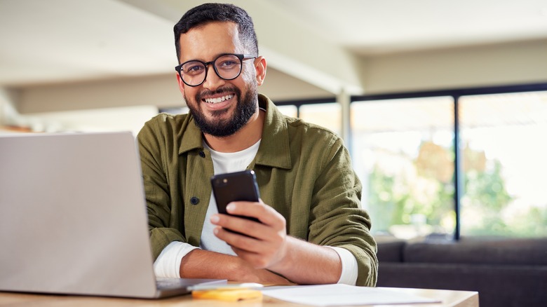 Man at desk smiling with phone in hand and laptop nearby.