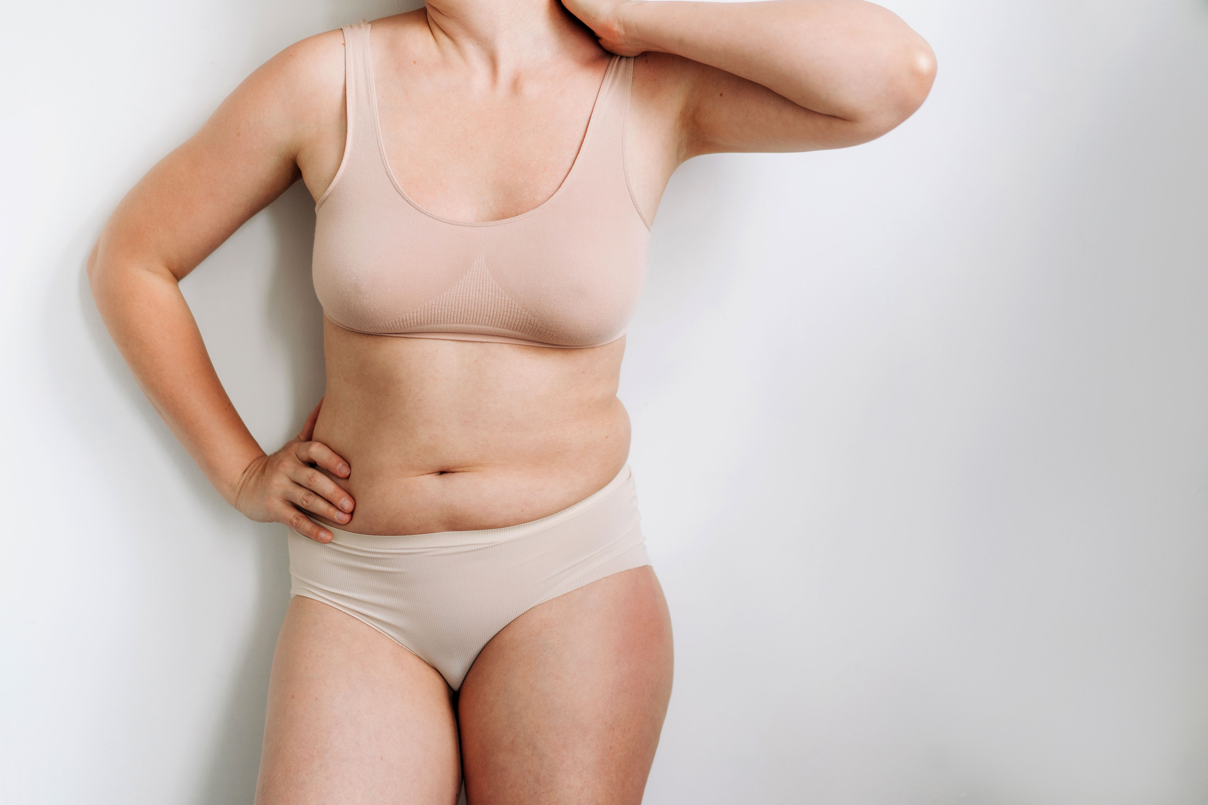 Person in neutral-toned underwear posed against a plain background, hand on hip, looking away from the camera