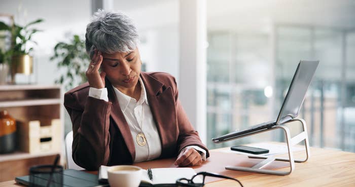 A person sits at a desk with a laptop stand, holding their head in apparent frustration, possibly overwhelmed by work