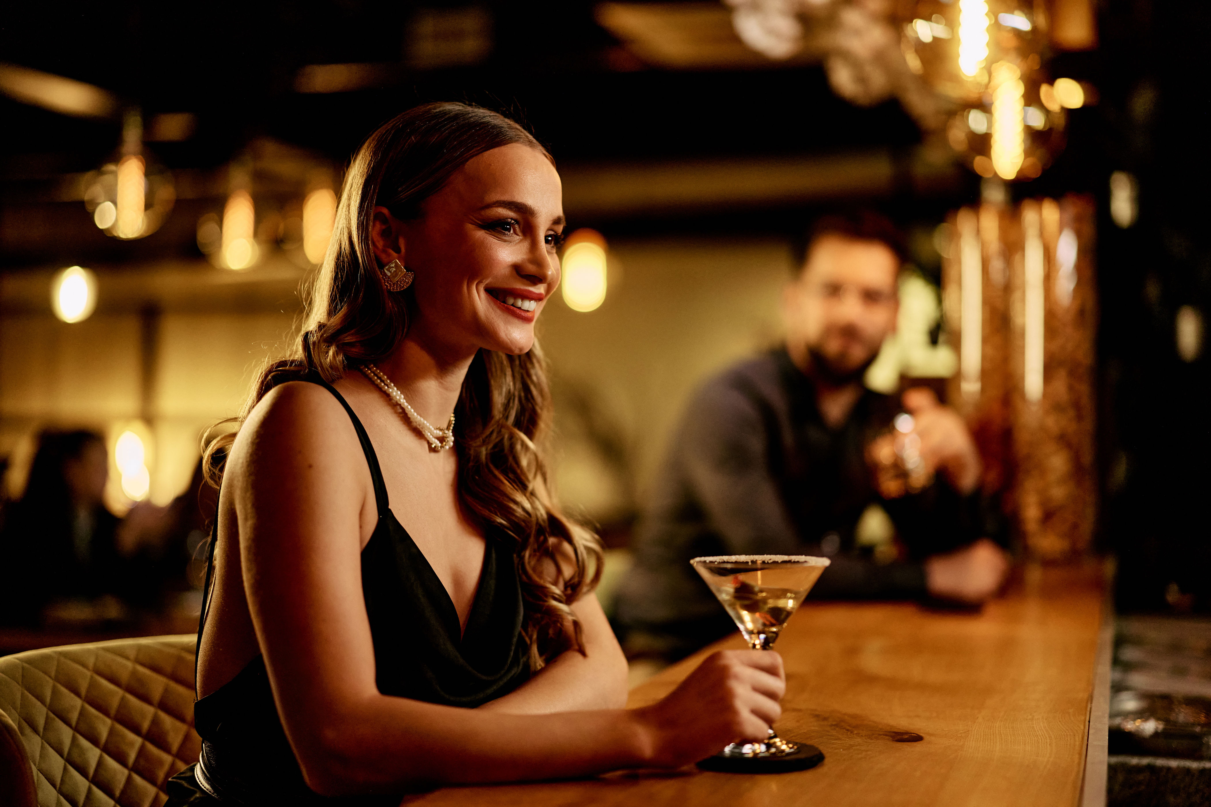 A person at a bar smiles while holding a martini glass, with another person in the background. The setting appears elegant and cozy