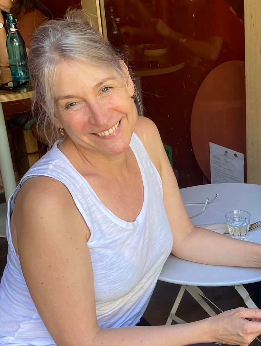 Person smiling while seated at a café table, wearing a sleeveless top and holding a glass