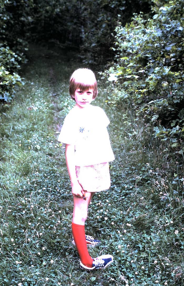 Child standing on a grassy path in shorts and a t-shirt, wearing one red sock and sneakers, surrounded by dense foliage