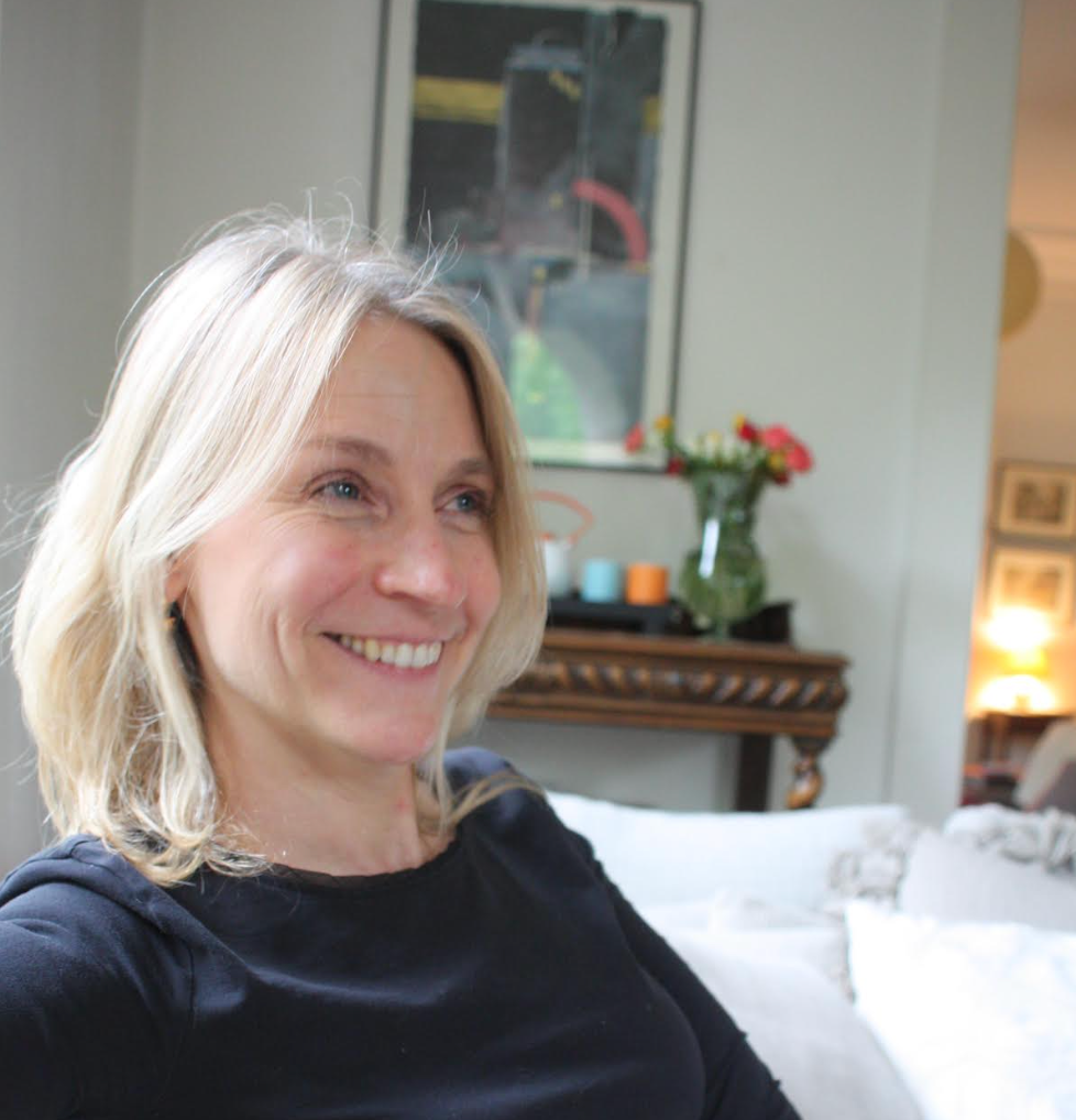 A woman with shoulder-length hair sits on a sofa, smiling, with a shelf and vase of flowers in the background