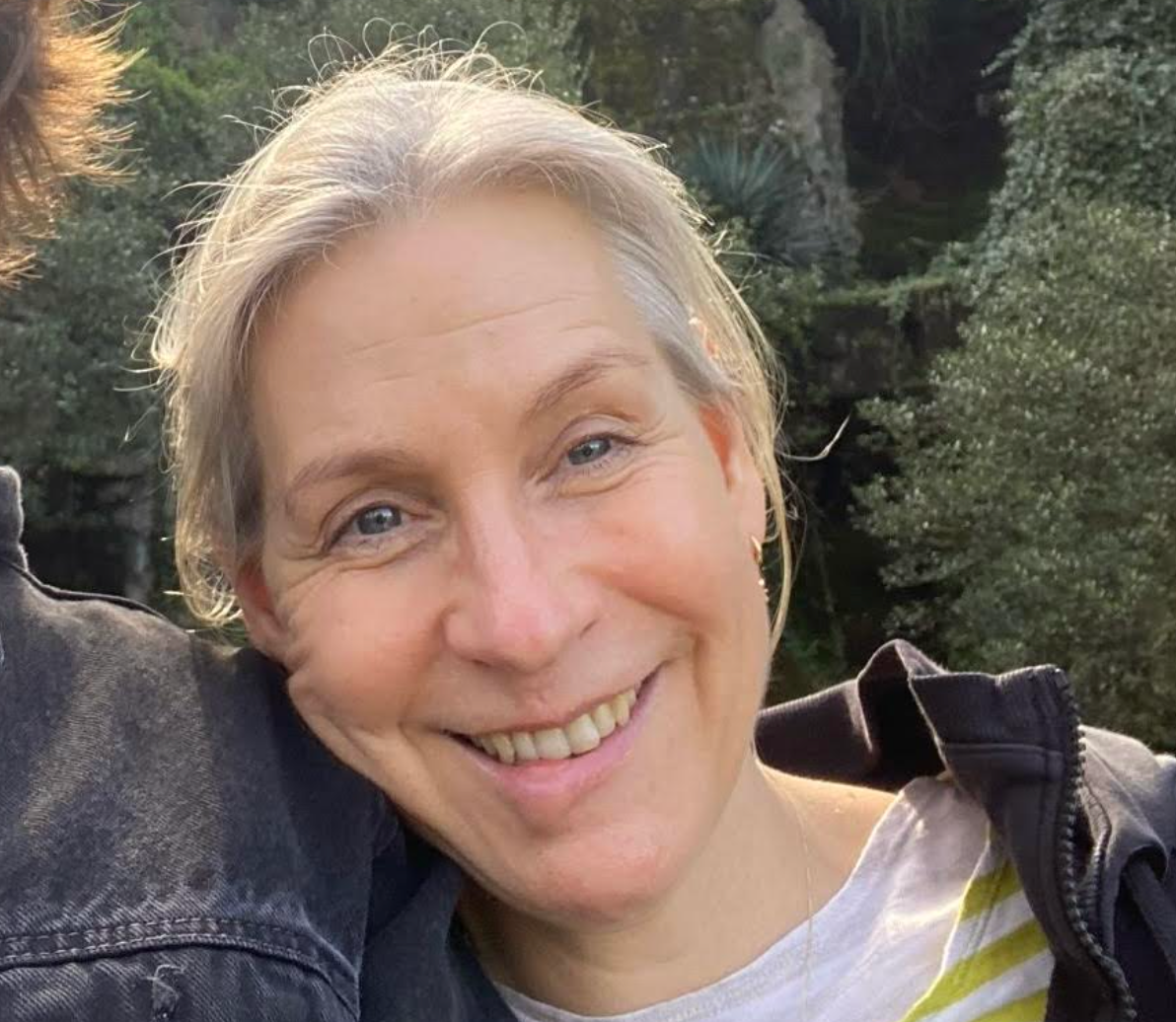 A person outdoors, smiling, wearing a light top and a jacket, with trees in the background