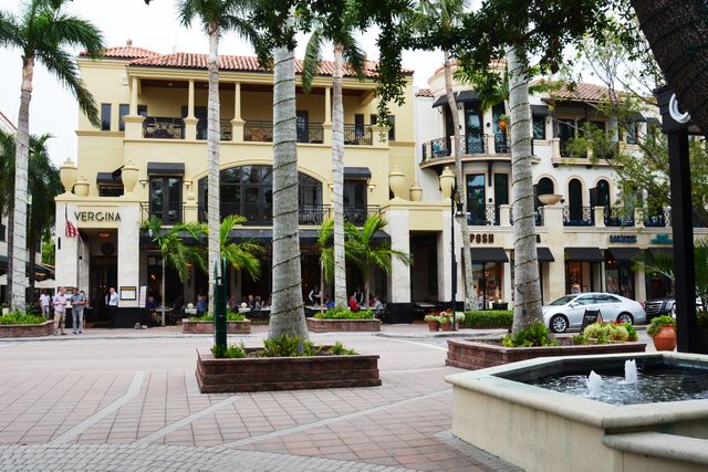 A commercial plaza with palm trees a fountain in the foreground and Mediterraneanstyle buildings housing various stores and restaurants