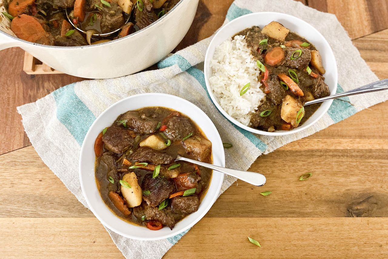Two Bowls of Jamaican Beef Stew (One Served With Rice and Just a Bowl of Beef Stew) on a Blue and White Kitchen Towel Next to a Dutch Oven With More Stew