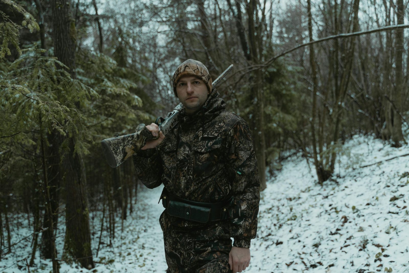 A man wearing camouflage gear holds a rifle in a snowy forest, showing a winter hunting scene.