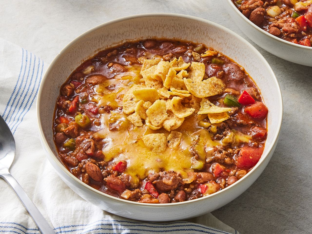 A bowl of chili topped with melted cheese and corn chips placed on a white cloth with a spoon nearby