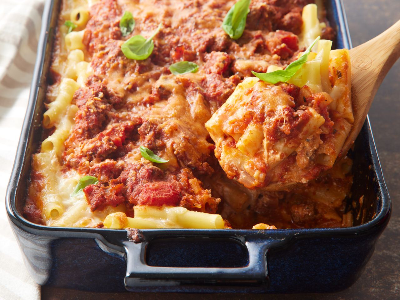 A baking dish of baked ziti, topped with fresh basil, with a wooden spoon holding up a big serving of baked ziti