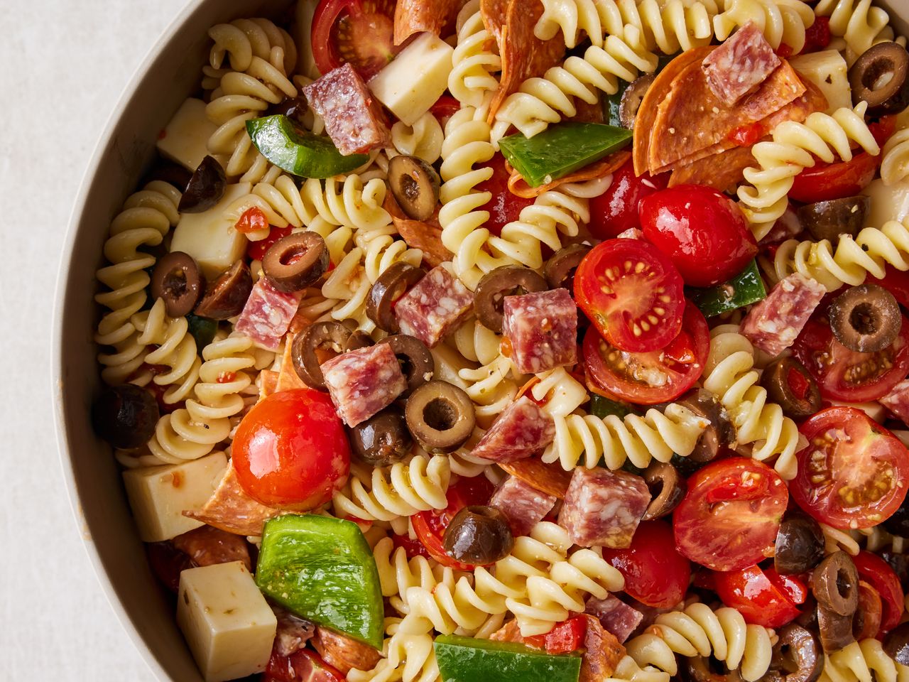 close up on a large bowl of pasta salad, with pasta, tomatoes, salami, pepperoni, peppers and cheese