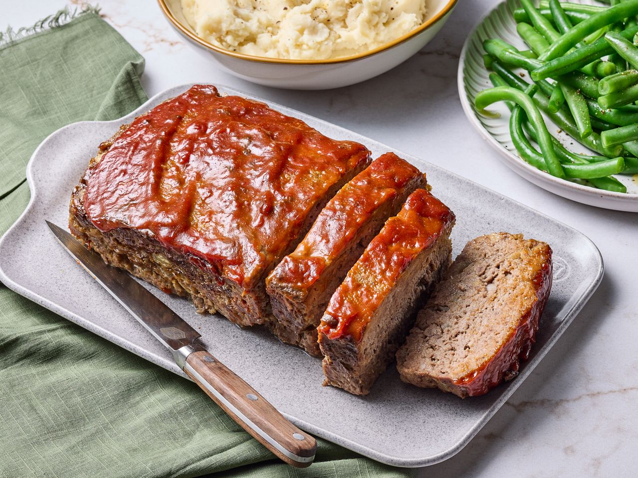Sliced glazed meatloaf on a platter with green beans and mashed potatoes in the background
