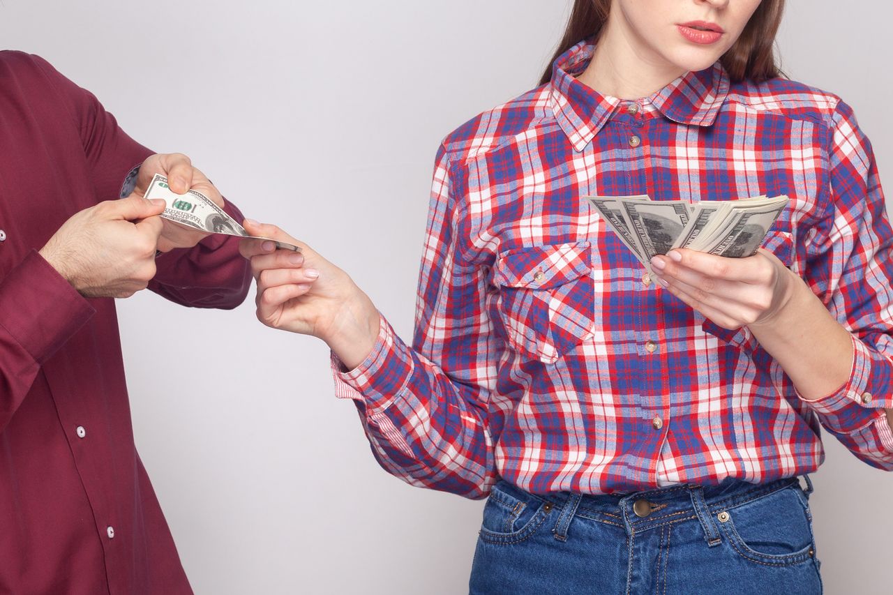 Portrait of serious rich confident woman and man gigolo standing together, wife giving money to her husband with indifferent face. Indoor studio shot isolated on gray background.