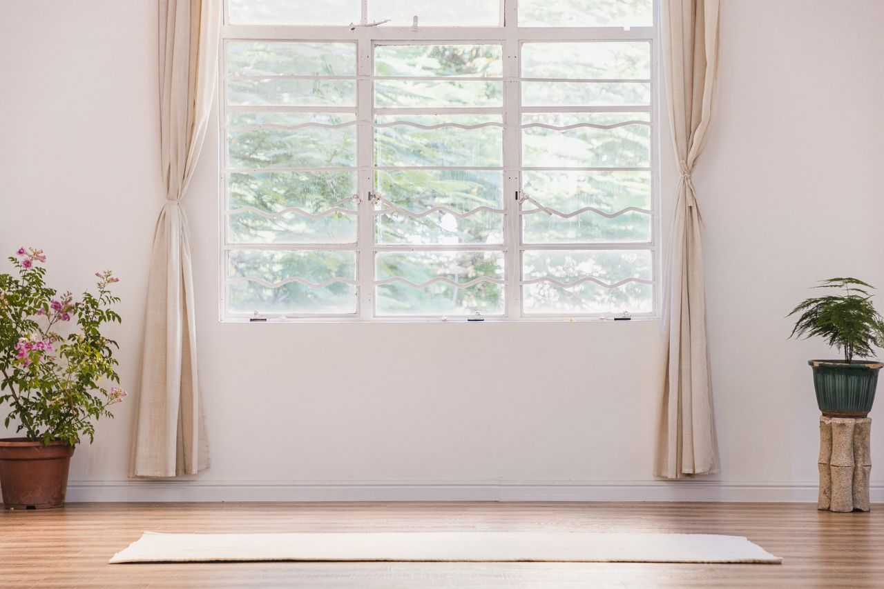 A minimalistic room with a mat on the floor a large window with curtains and potted plants arranged on either side