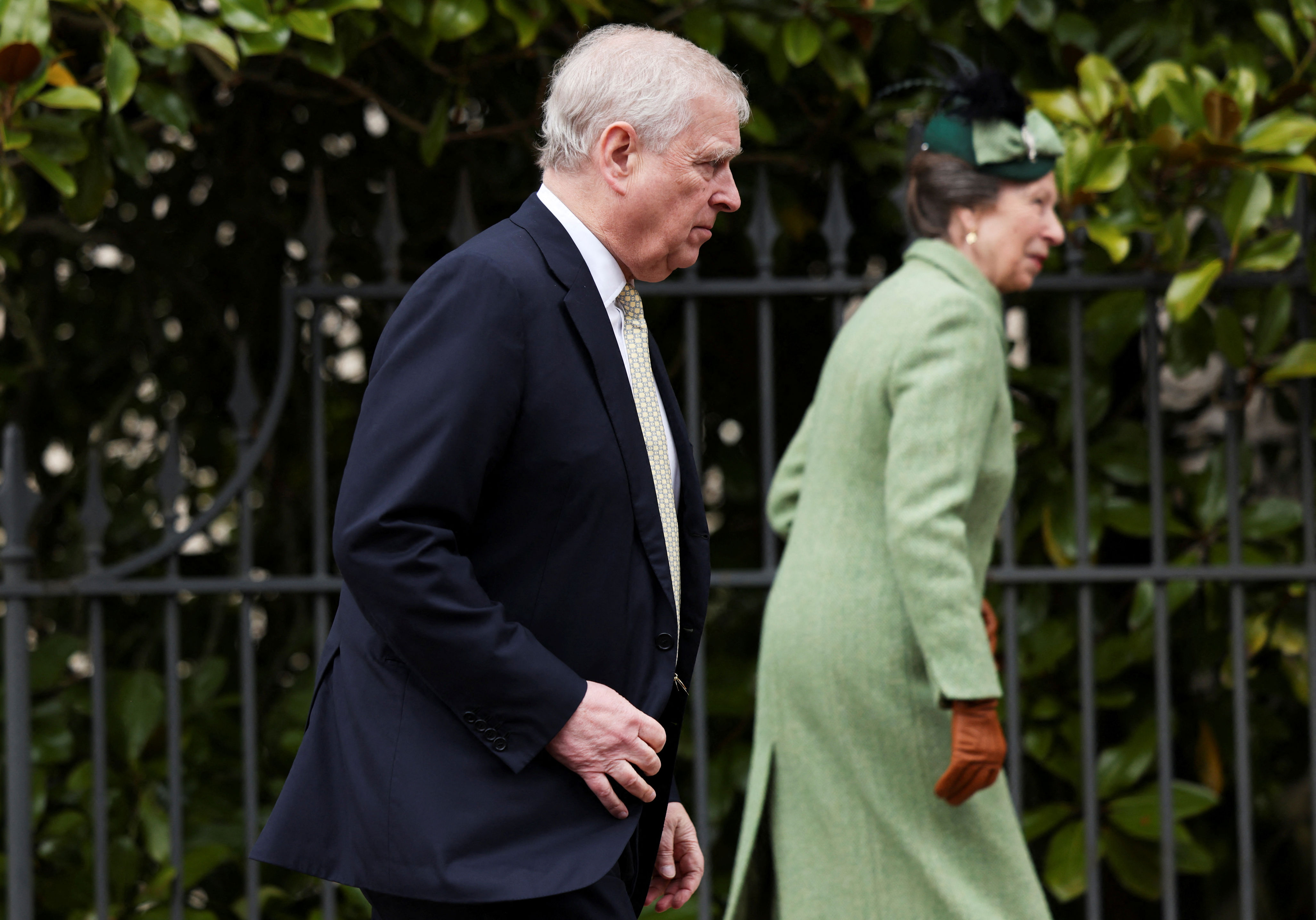 Prince Andrew walking behind Princess Anne walking by a fence with trees