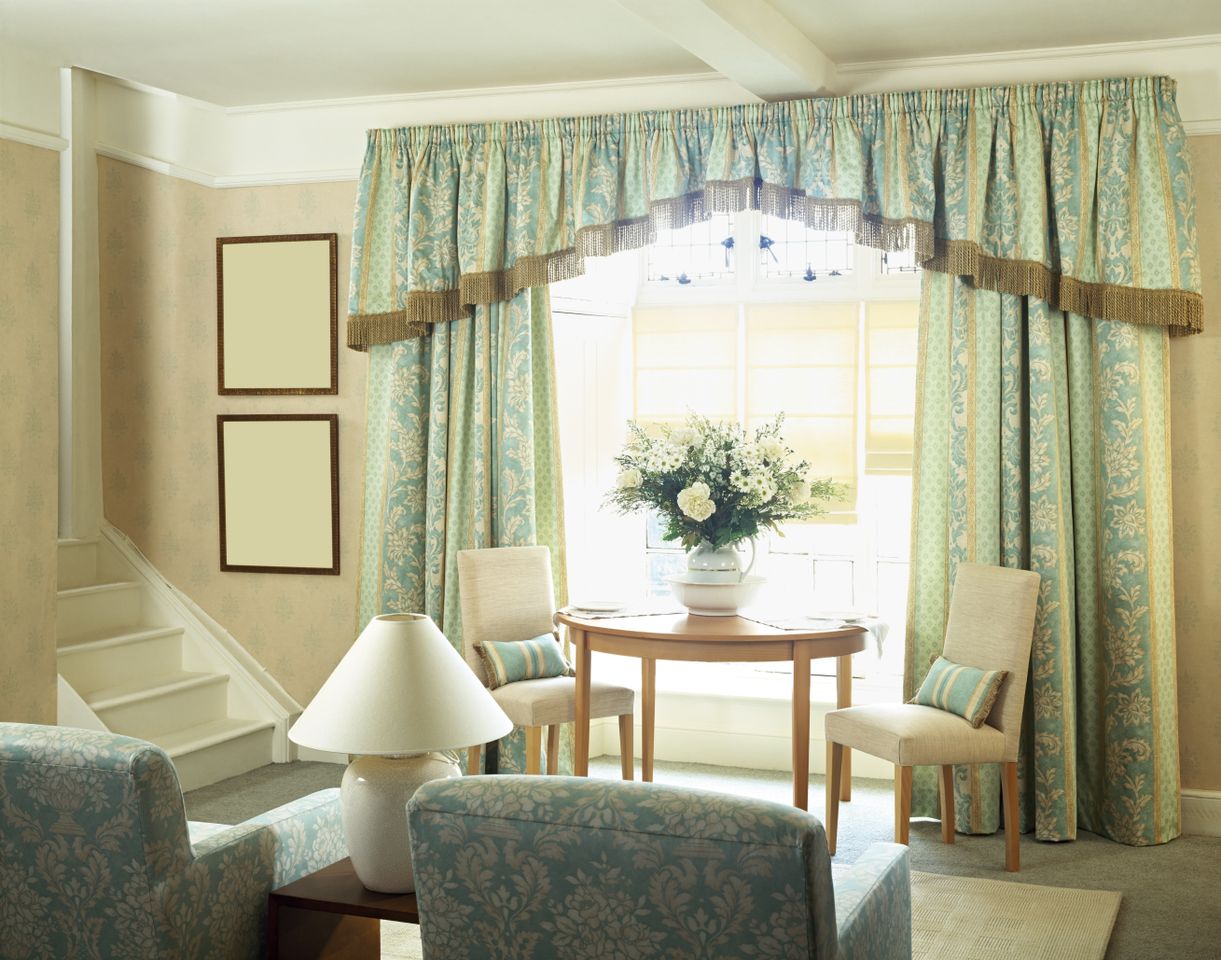 A living room with a table and flower arrangement in front of a window with curtains two upholstered chairs and a staircase to the side