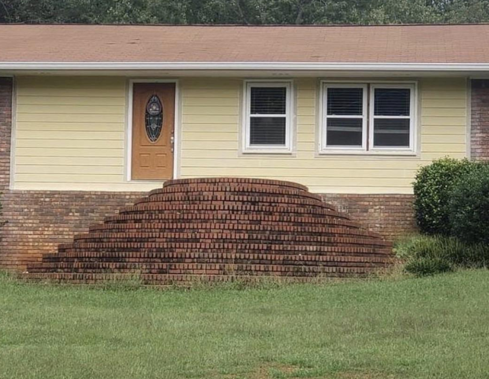 House façade with a yellow door and windows. Unusual semi-circular brick staircase leads to the door, causing a sloped design. Bush on the right