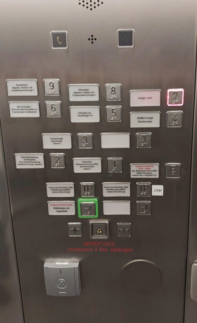 Elevator control panel with numbered buttons, emergency call button, and multiple floor labels in German. The 7th floor button is lit