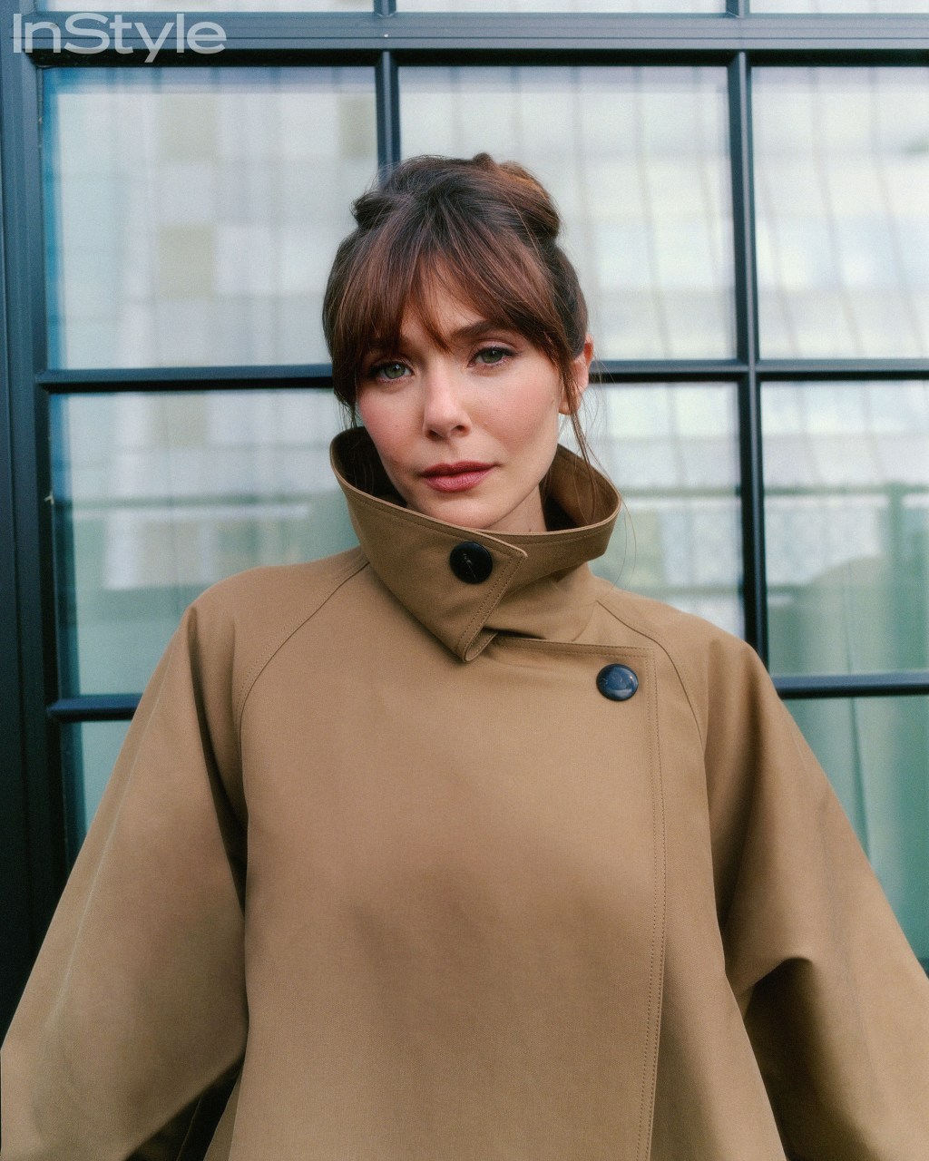 Elizabeth Olsen wearing a tan trench coat, looking directly at the viewer.