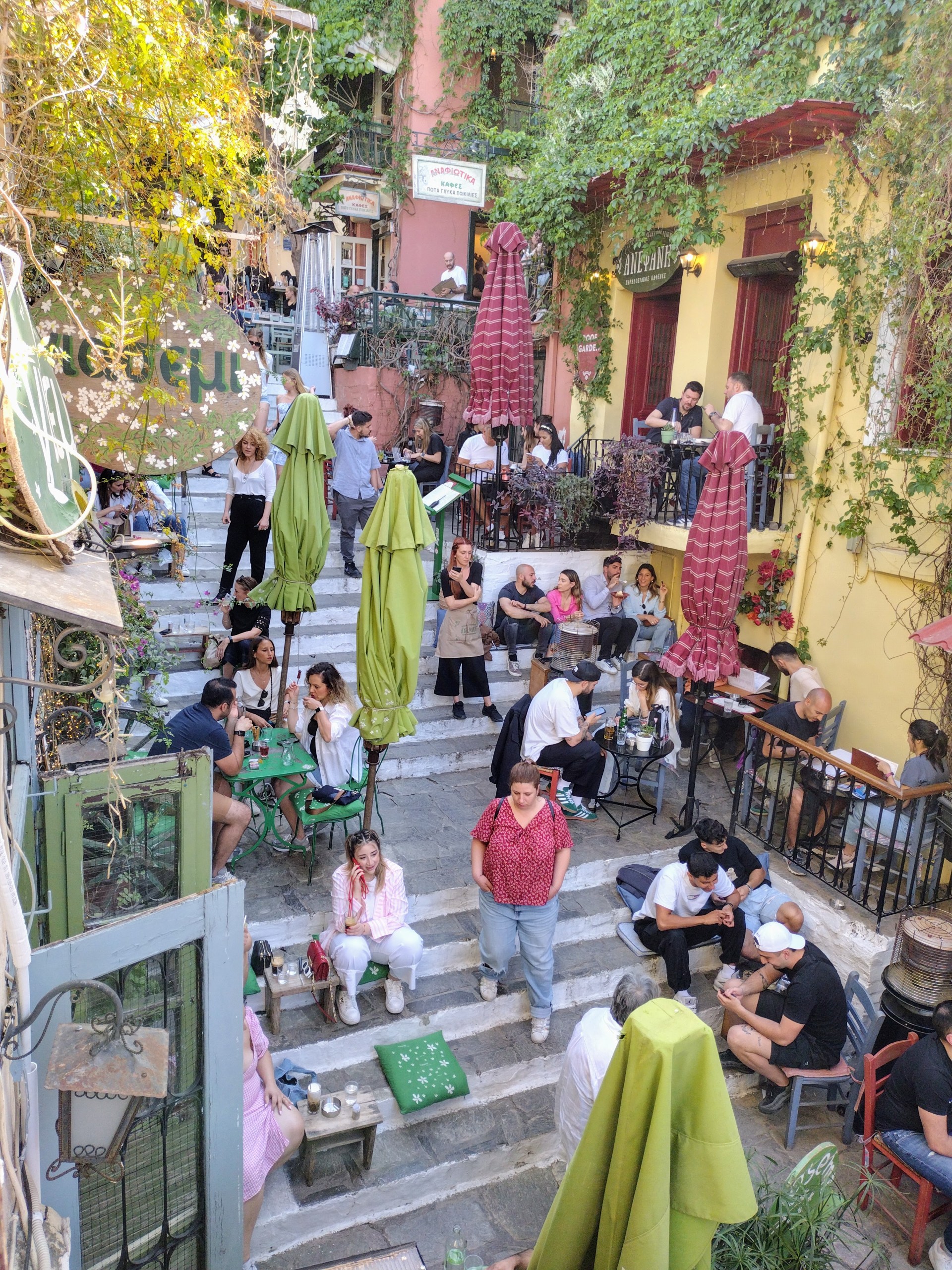 People eating at a restaurant split over steps on Mnisikleous Street, Athens. Vines drape down over the building and there are red umbrellas folded up.