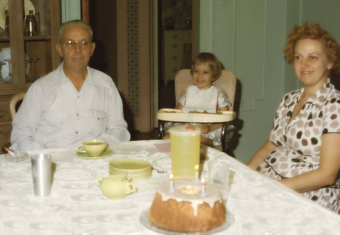 Older man, woman, and toddler smiling at a dining table with a cake and drinks. The setting is a home, suggesting a family celebration