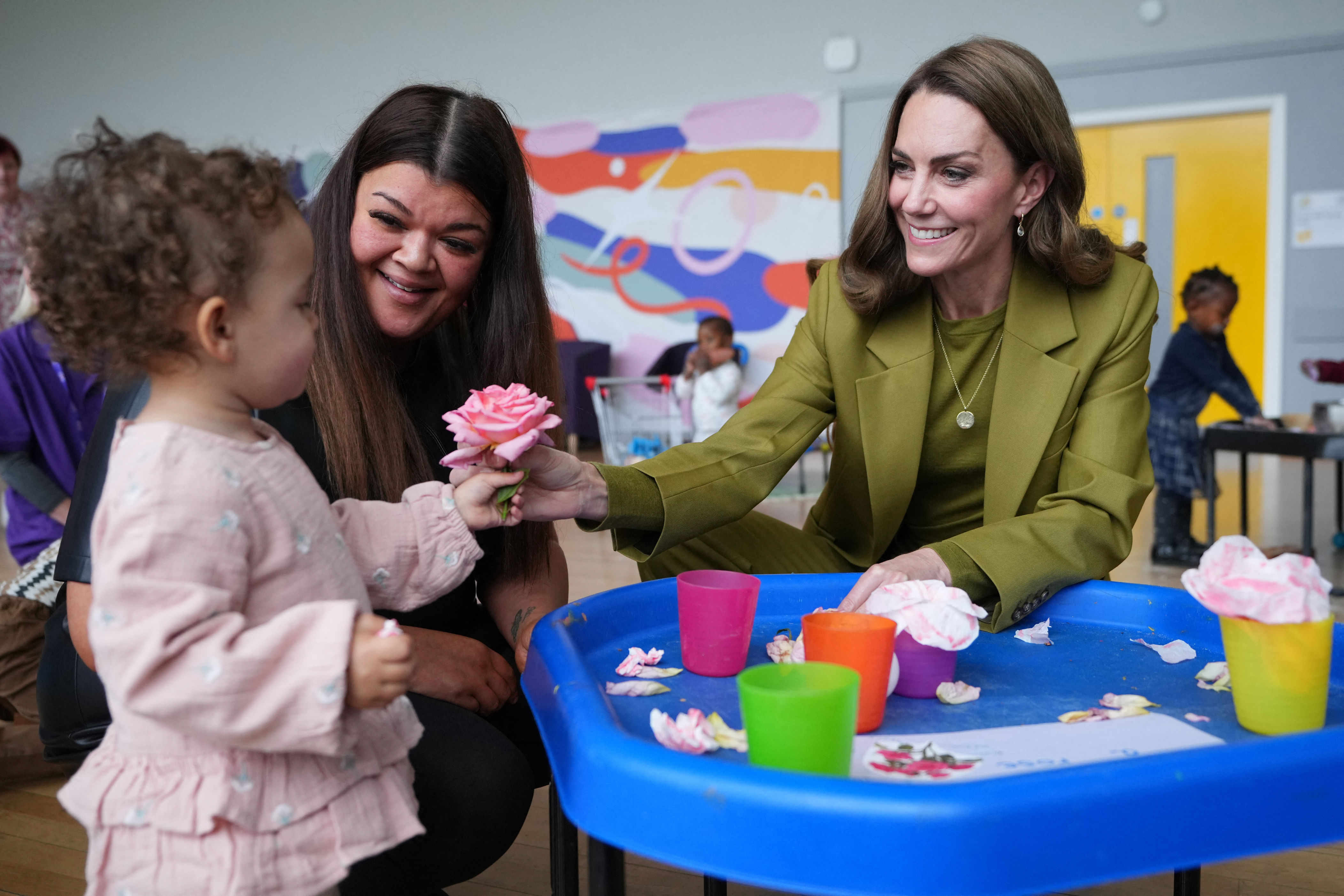Kate Middleton sitting at a table in a green suit handing a little girl a rose