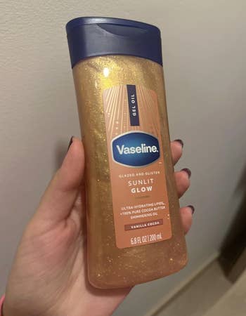 A hand holds a bottle of Vaseline Glazed and Glisten Sunlit Glow gel oil with shimmering particles, labeled as ultra-hydrating and vanilla cocoa scented