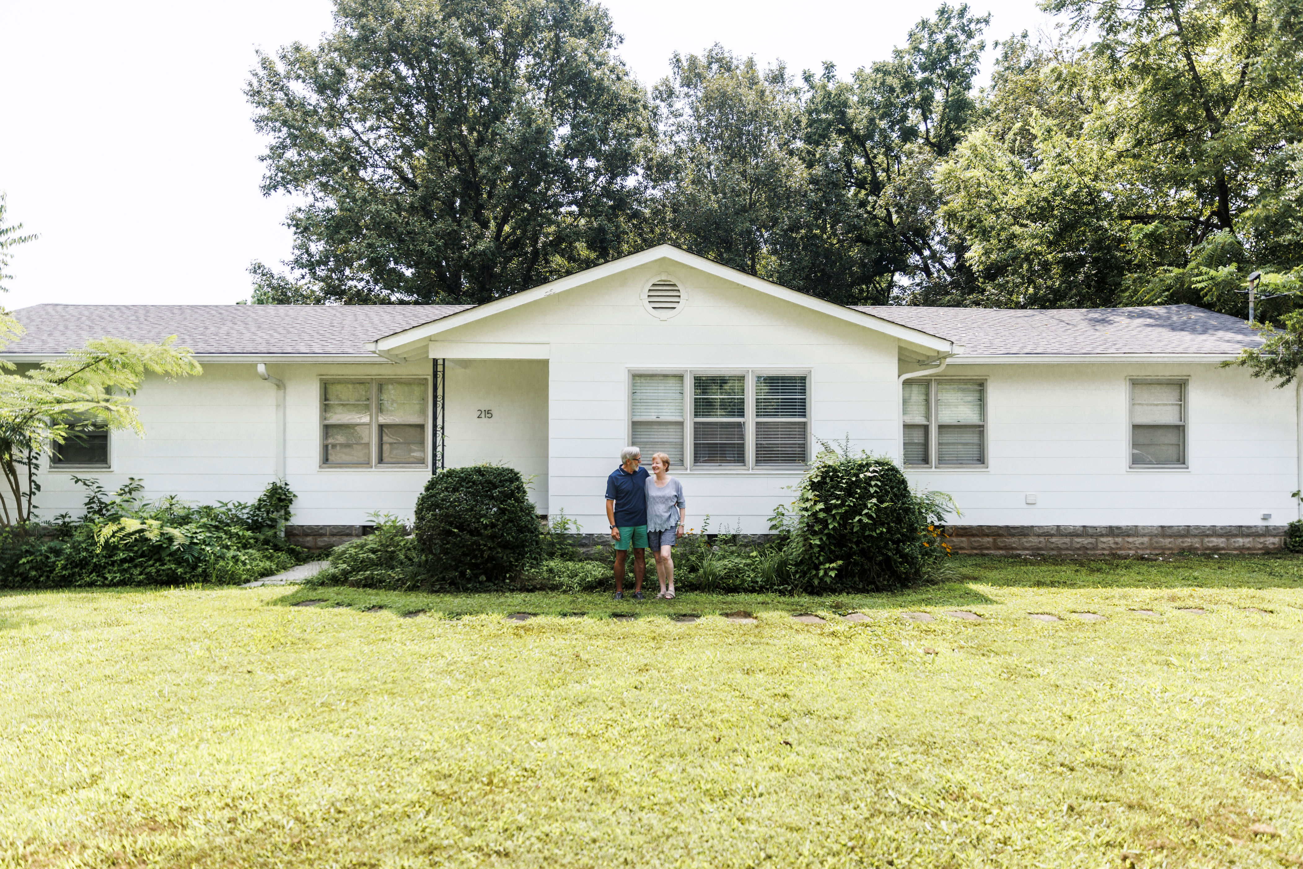 A couple stands in front of a suburban house, surrounded by greenery and trees, on a sunny day