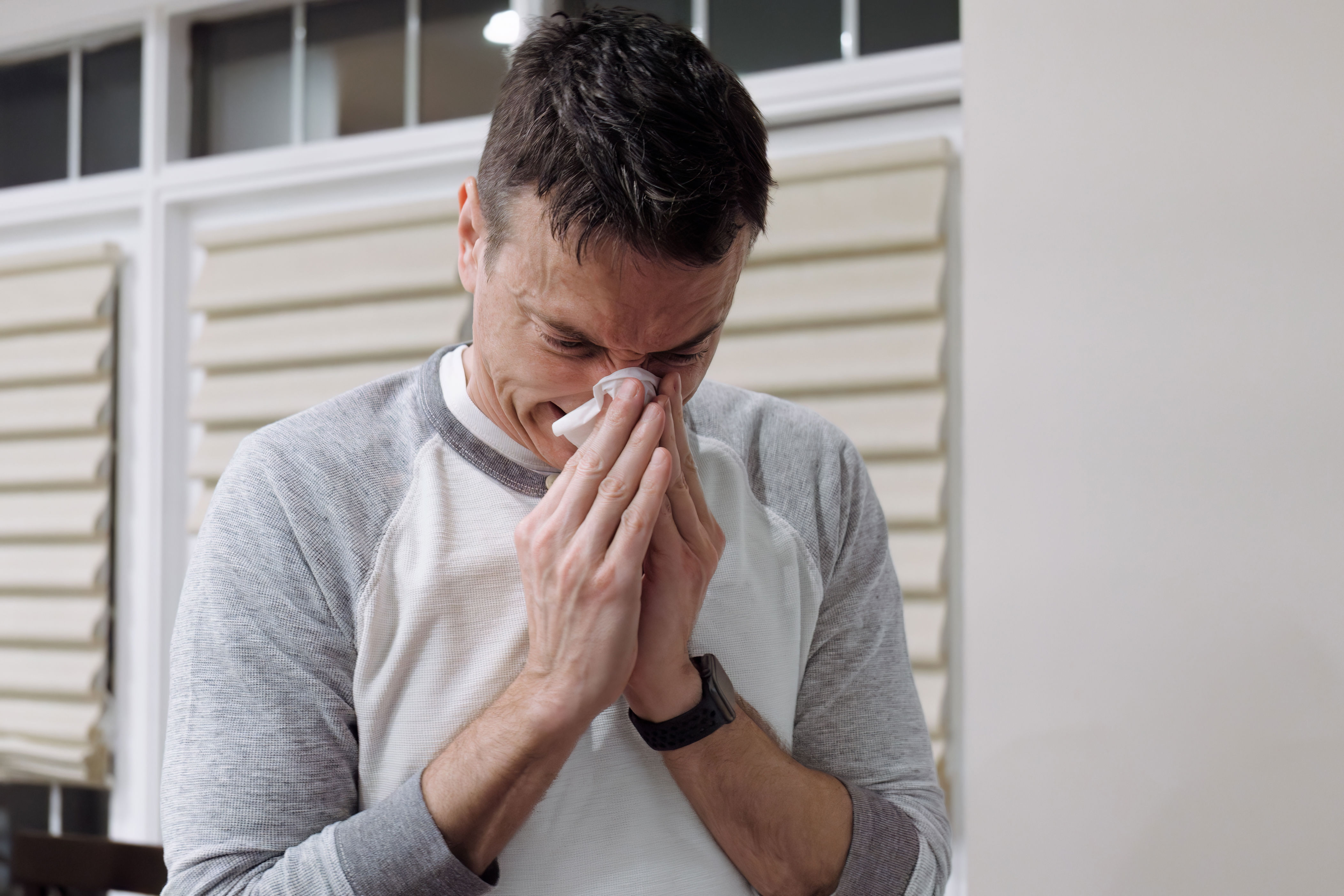 Person with short hair wearing a casual two-tone shirt, standing indoors, appears to be sneezing or wiping their nose with a tissue