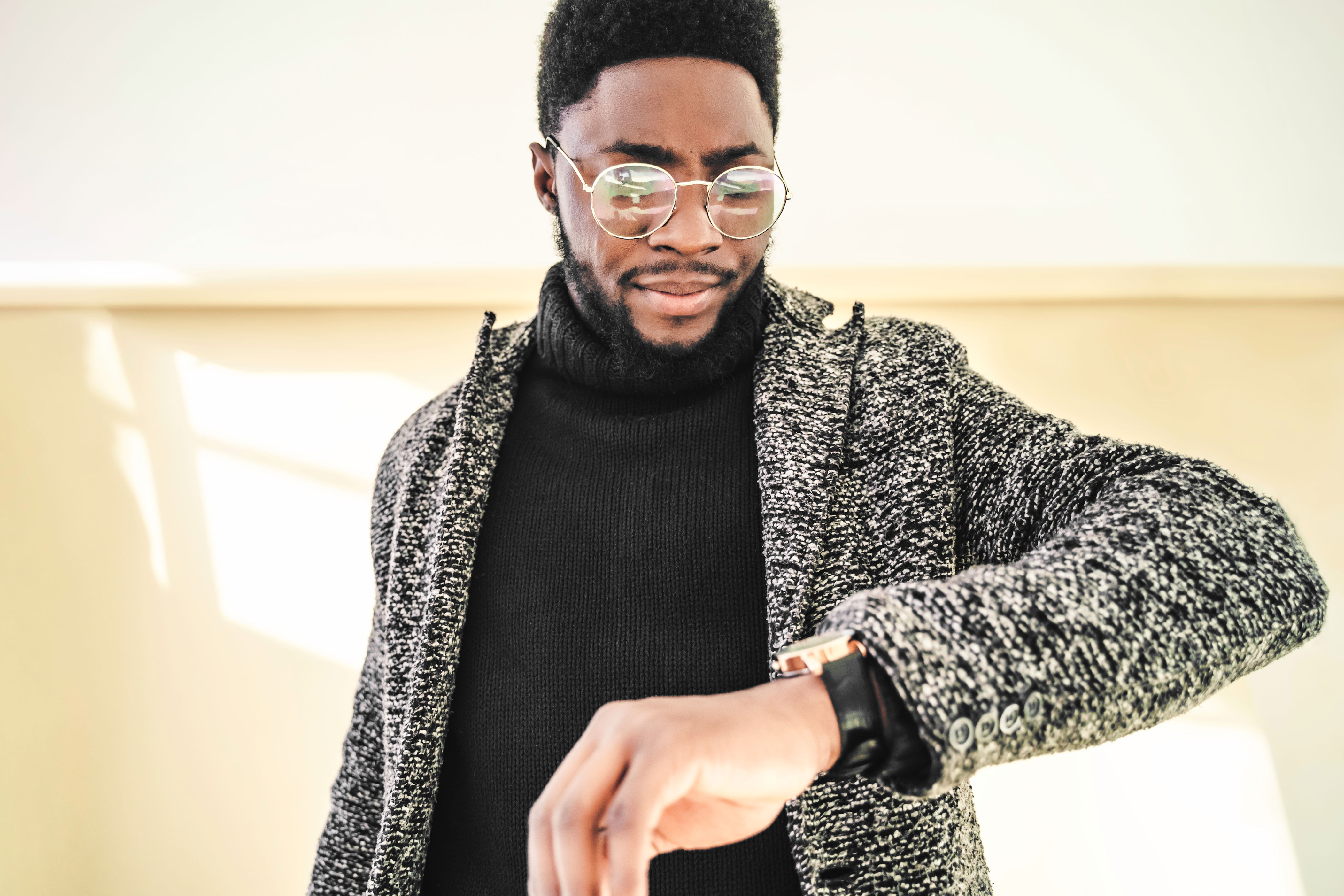 Man in a textured jacket and glasses checking a watch on his wrist, standing indoors with a neutral background