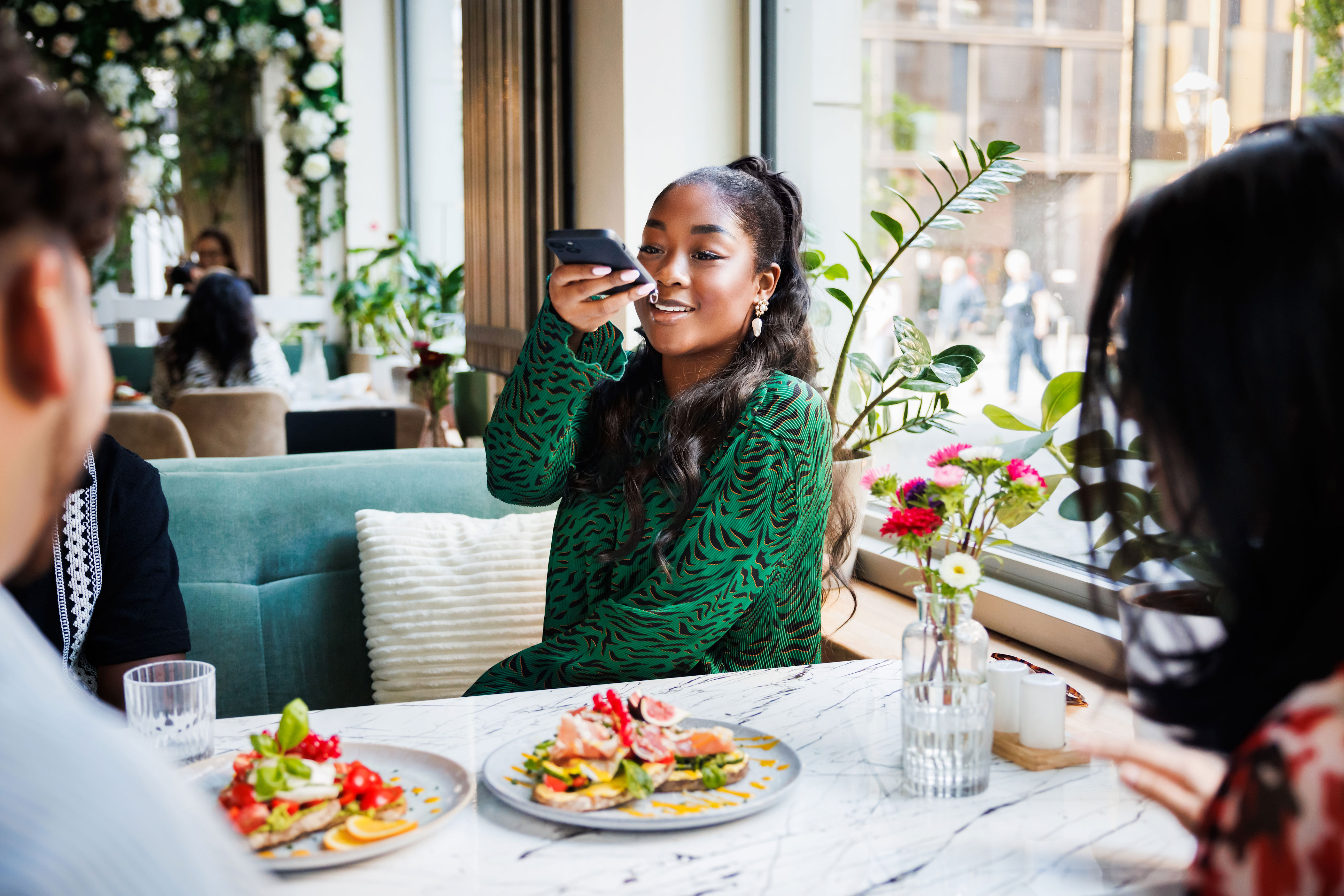 Person in a patterned shirt takes a smartphone photo of food at a restaurant table with friends