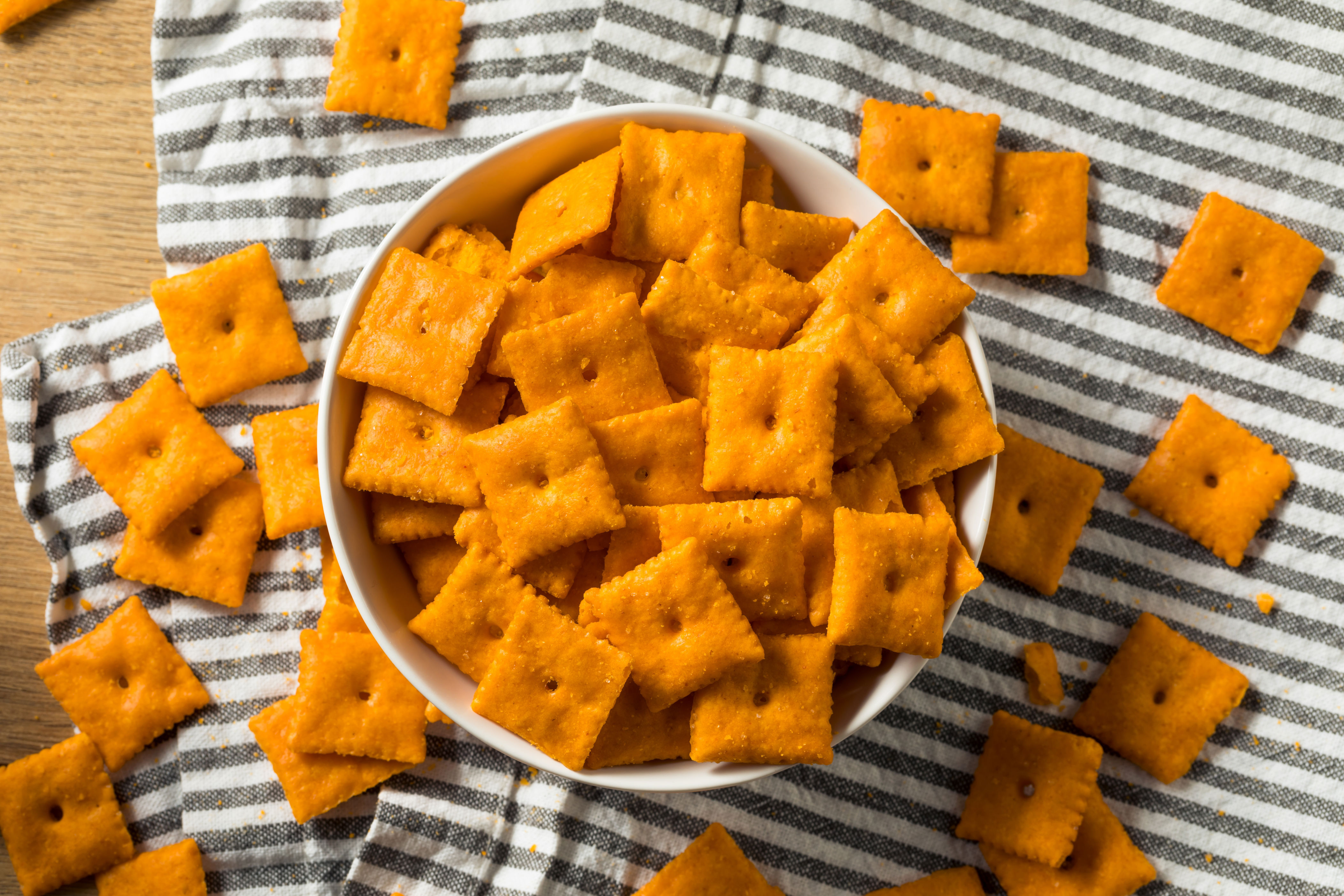A bowl filled with square cheese crackers sitting on a striped cloth, with some crackers scattered around the bowl