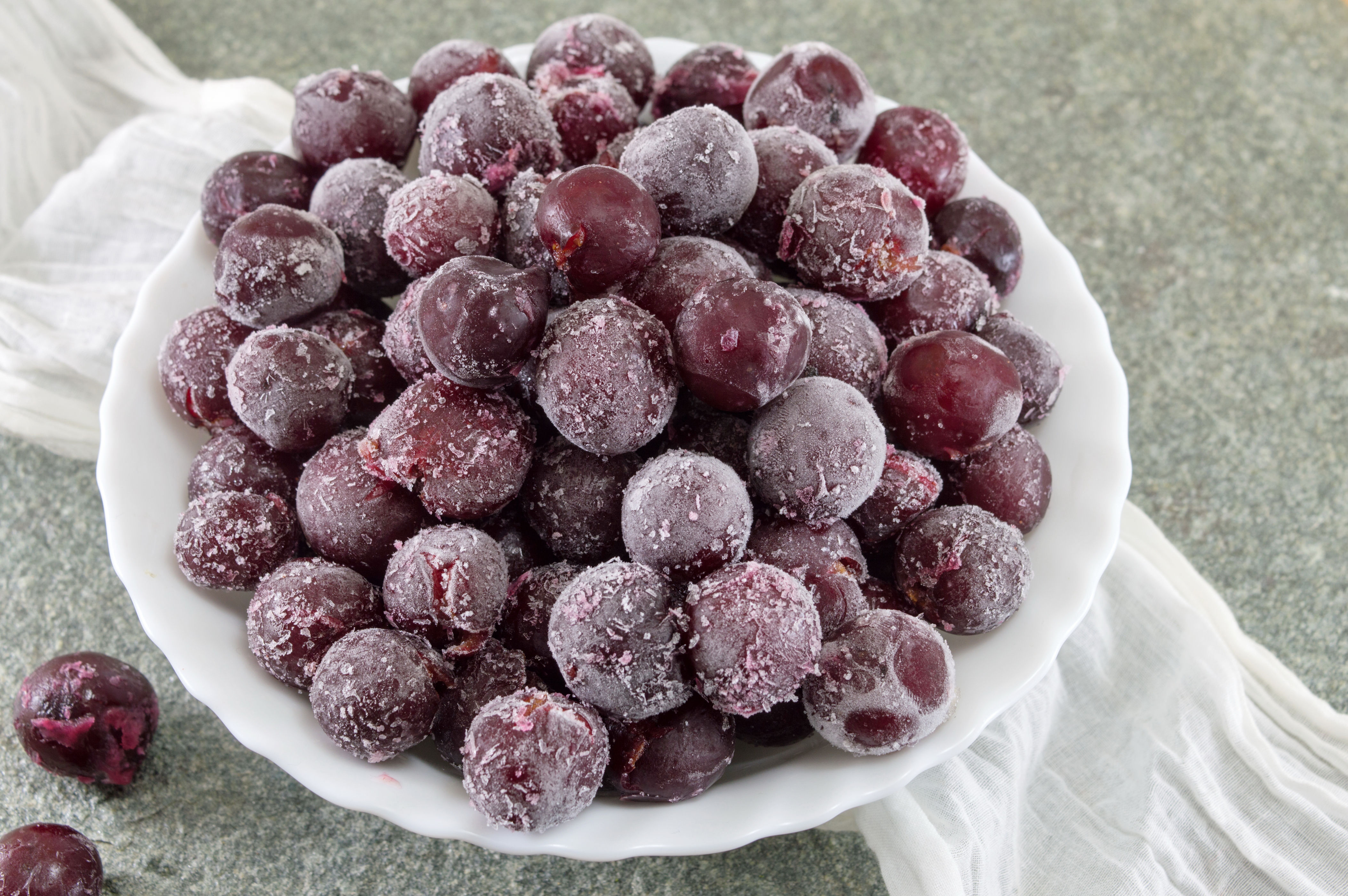 Frozen cherries piled in a white dish on a textured surface, with a white cloth beside them