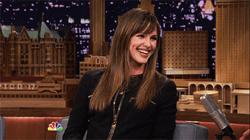 jennifer garner smiling and wearing a zip-up jacket, sitting on a talk show set with a cityscape backdrop and microphone