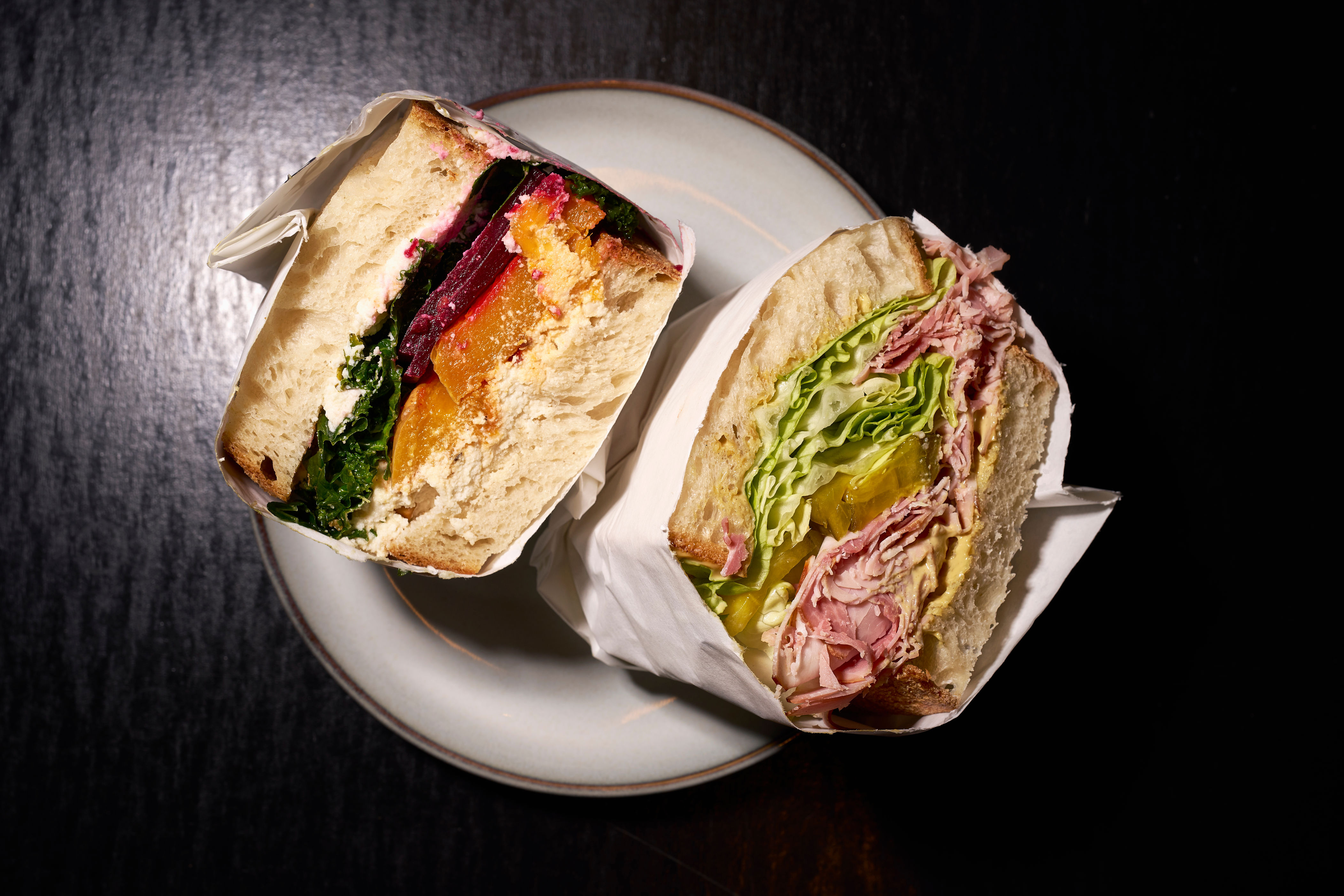 Two halved sandwiches on a plate: one with roasted vegetables and hummus, the other with ham, lettuce, and mustard