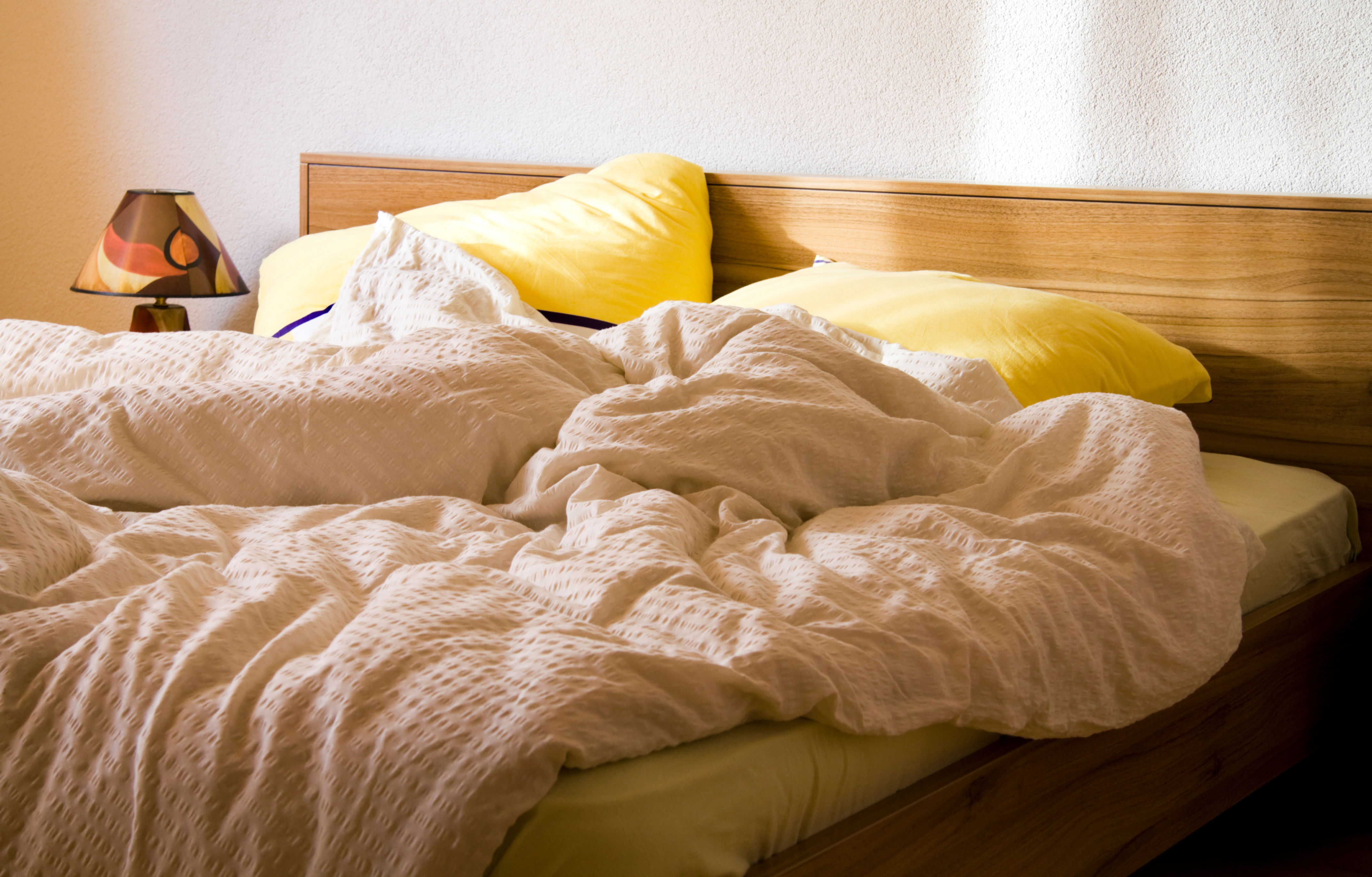 Unmade bed with crumpled sheets and pillows, beside a small lamp on a wooden nightstand