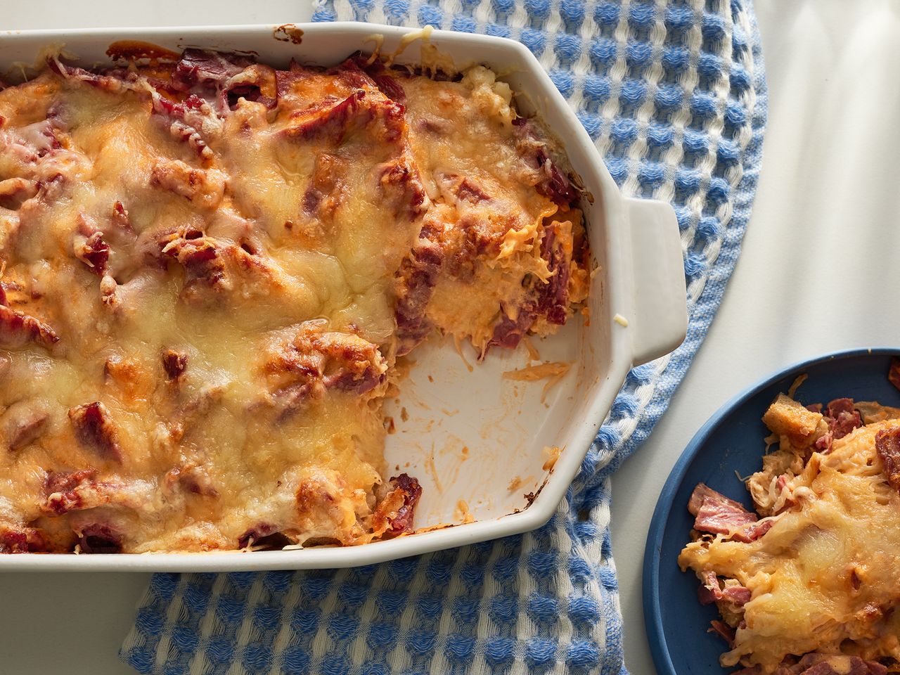 close up view of a Reuben Casserole in a white baking dish and a slice of Reuben Casserole on a blue plate