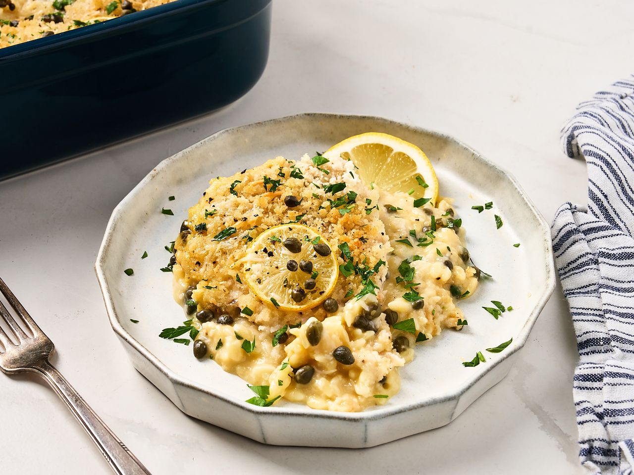 A plate of chicken piccata casserole with sauce breadcrumbs capers and a lemon slice garnished with herbs