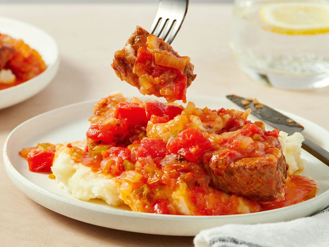 A plate of Swiss steak served with a tomatobased sauce over mashed potatoes a fork lifting a piece of the steak
