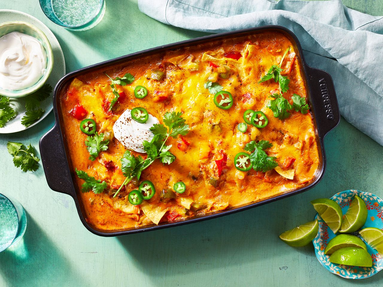 looking down at a casserole dish of king ranch chicken casserole, garnished with fresh herbs, pepper slices and sour cream, next to a plate with lime wedges