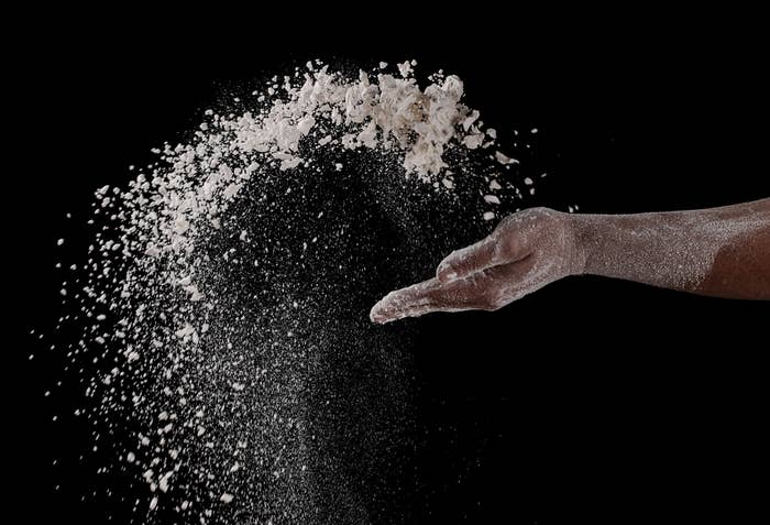 A hand gracefully tosses flour in the air against a black background, emphasizing movement and texture