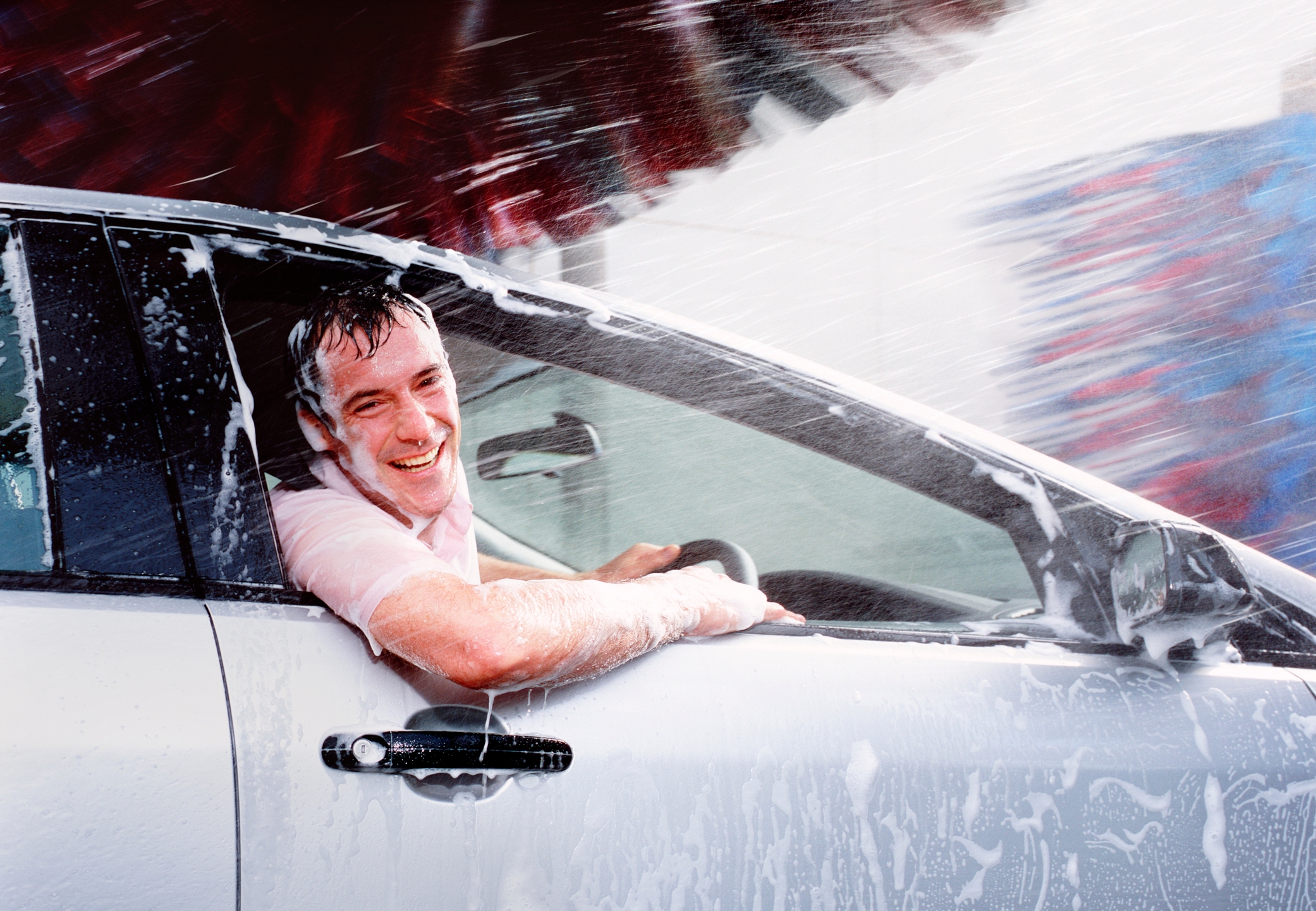 Person in a car wash, smiling with a soapy car and wet hair, leaning out the window as brushes spin in the background