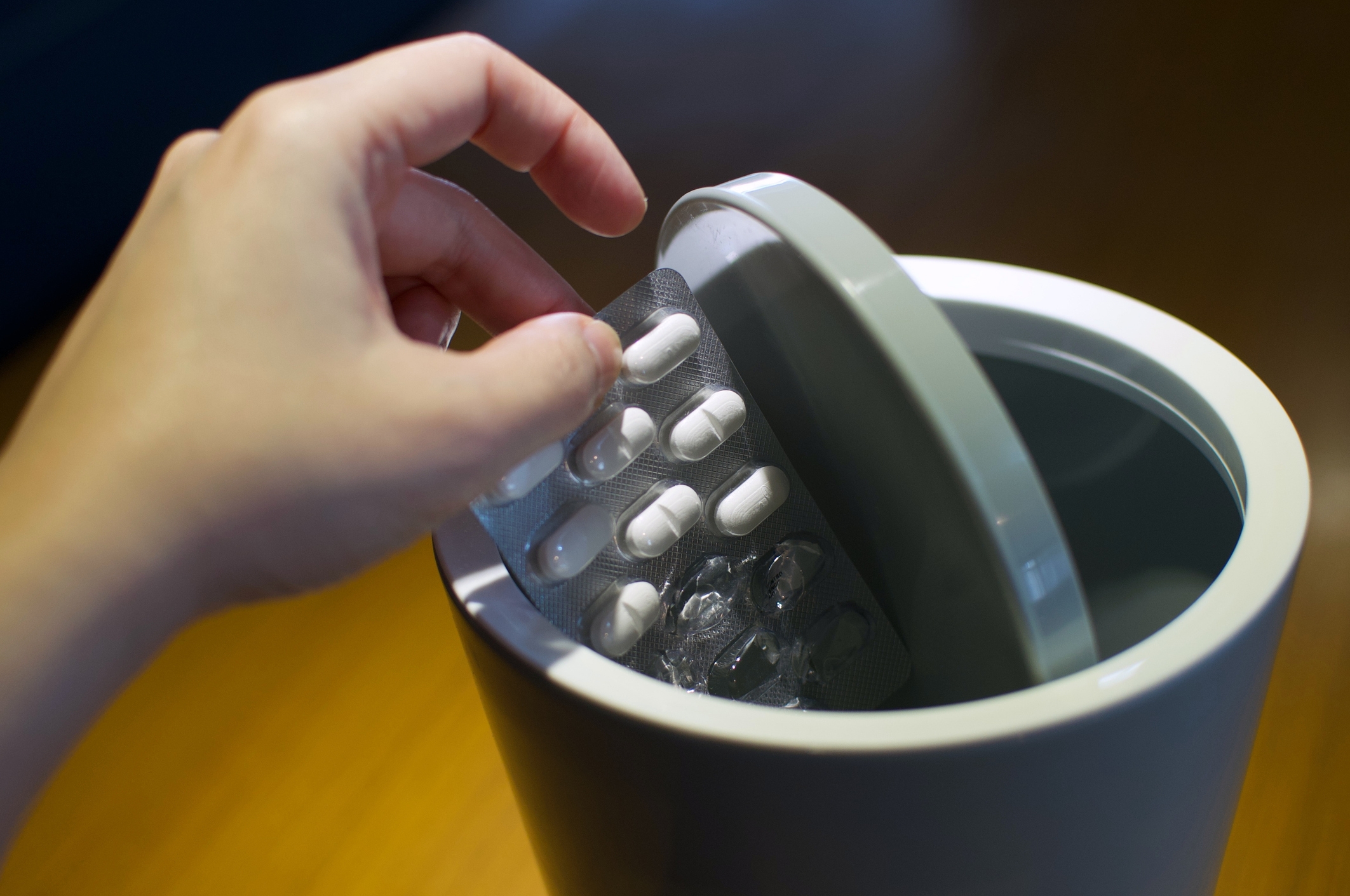 A hand is seen discarding a blister pack of pills into a trash bin, suggesting medication disposal